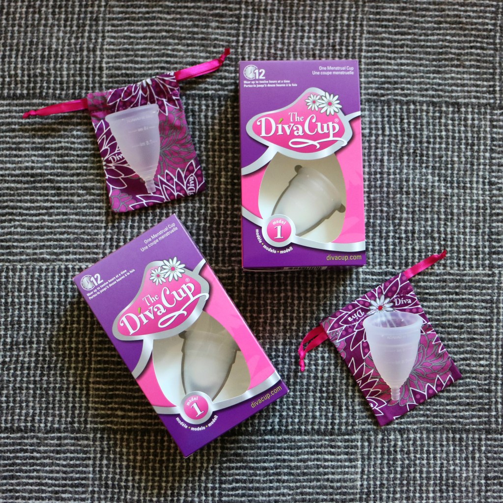 Four Diva Cups laid out on a grey woollen blanket. Two are in their purple and pink packaging while two are sitting on the purple floral pouches.
