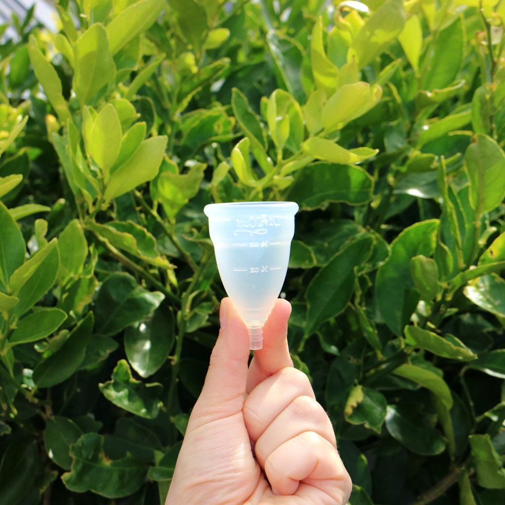 A single Diva Cup held in front of bright green foliage.